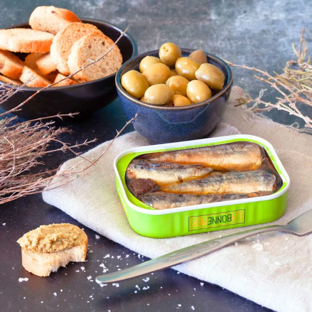 opened sardine tin with a bowl or olives and bread slices on a table