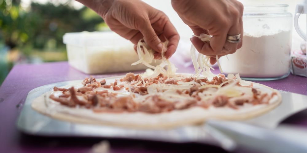 Hands topping a Flammekueche with onions and lardons before baking