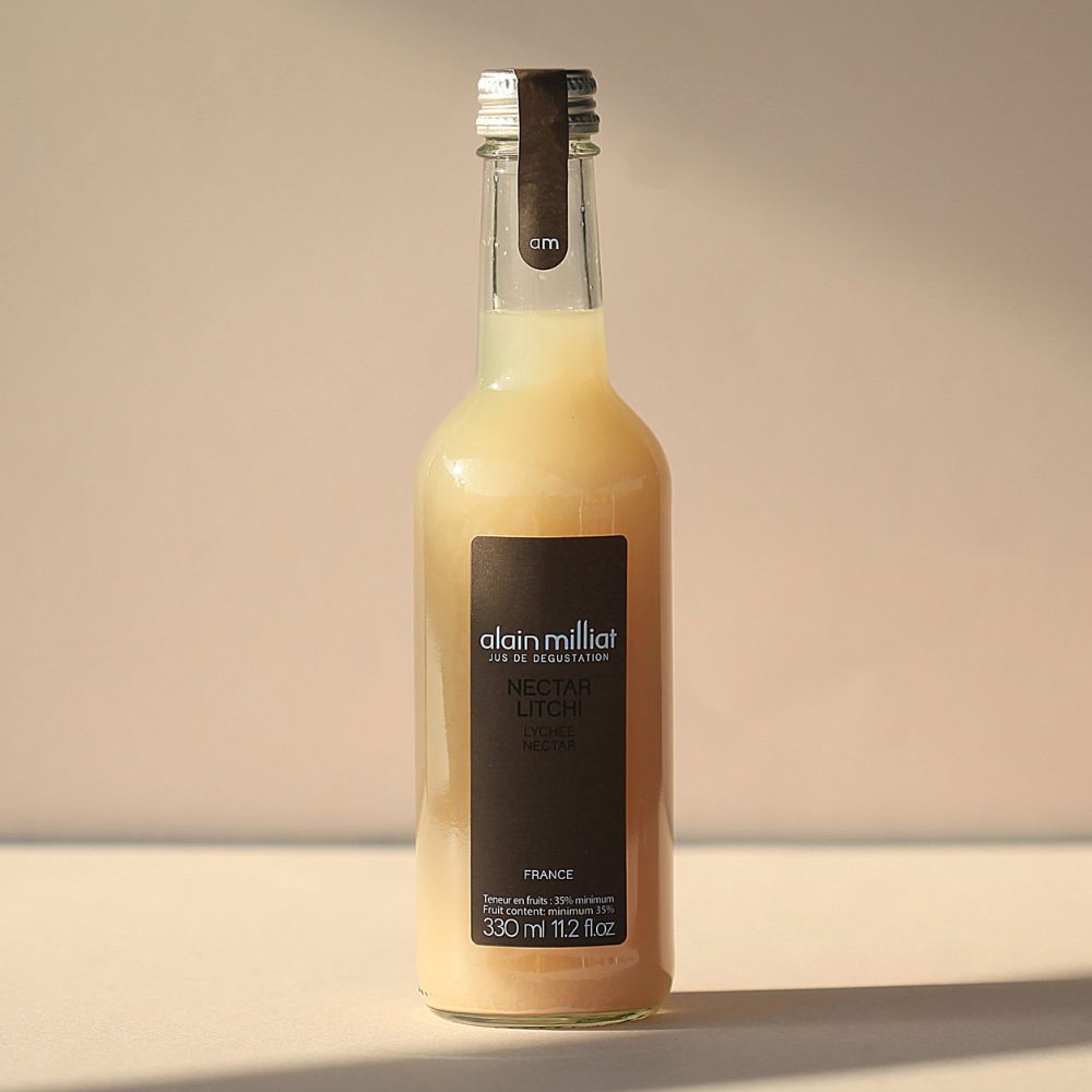 Bottle of Alain Milliat lychee nectar filled with pale, cloudy fruit juice, standing in warm sunlight against a soft beige background with a long shadow cast across the surface.