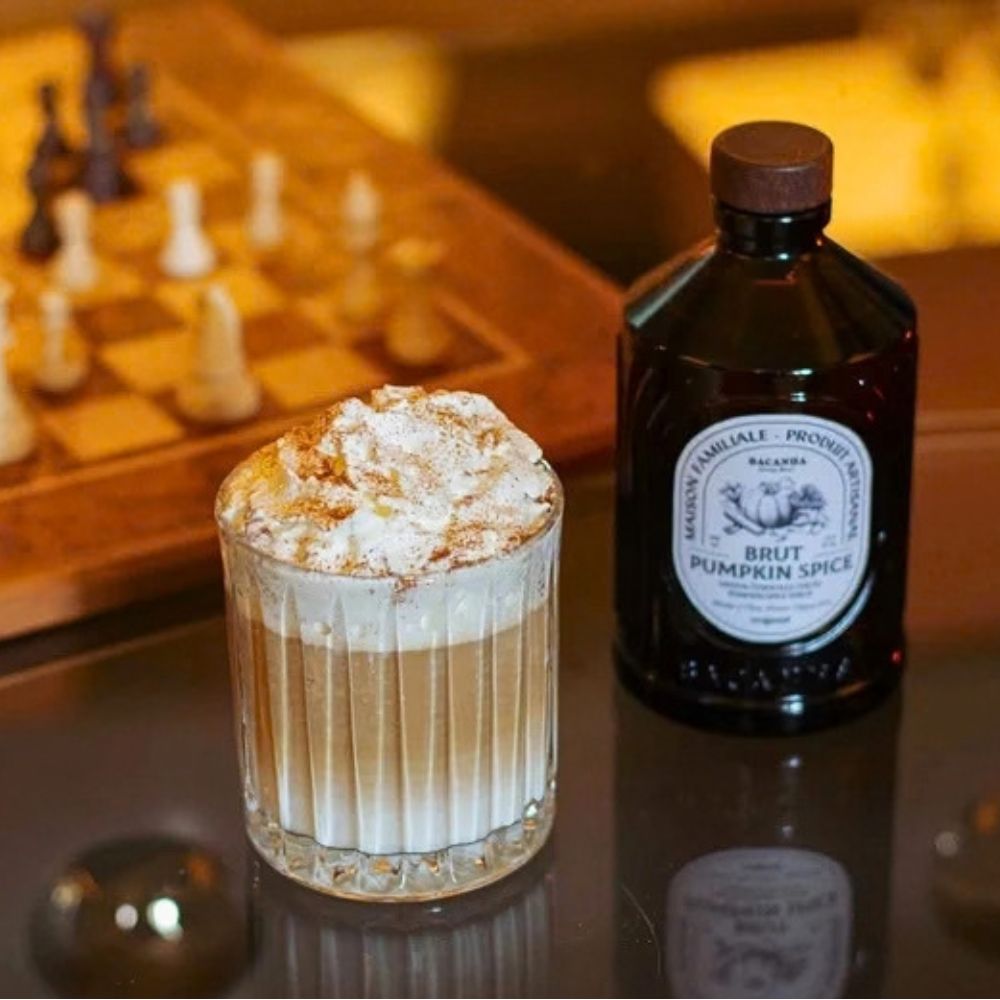 Bacanha Pumpkin Spice syrup bottle displayed beside a creamy, whipped-cream-topped spiced drink in a glass, photographed in a warm café setting.